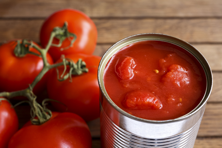 Open tin of chopped tomatoes with whole fresh unfocused tomatoes behind. Wood surface. Food service purchasing