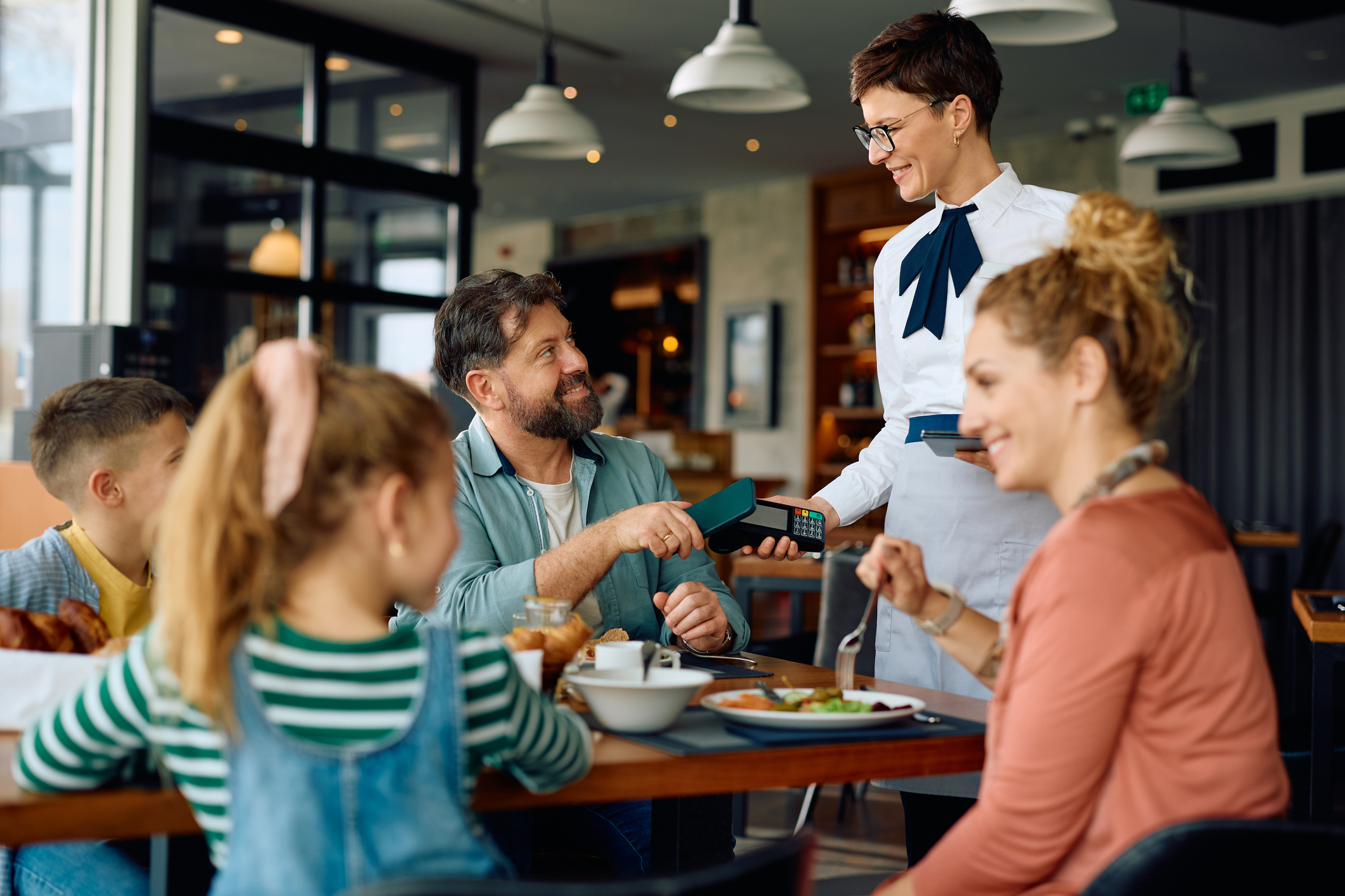 Happy father using mobile phone while paying to a waitress in restaurant at the hotel.