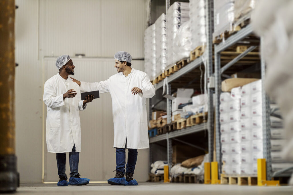 Two men interact joyfully at their job in a warehouse. They're wearing PPE, including hairnets, shoe covers, and jackets.